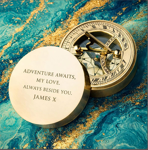 A brass nautical compass with a personalised engraving on its lid, set on a deep blue marbled background.