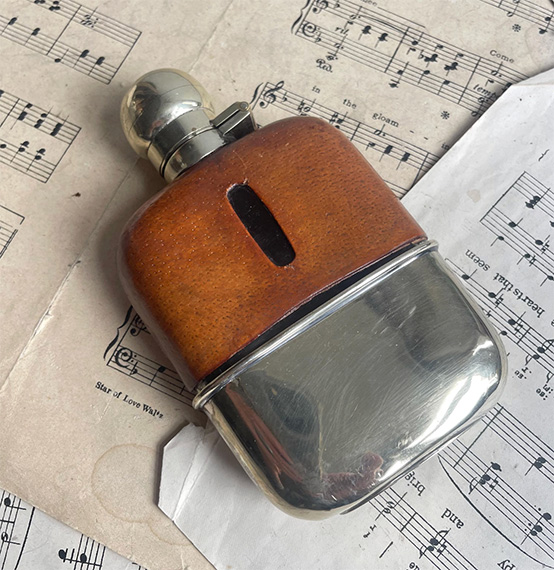 A vintage English leather-mounted glass hip flask resting on sheet music, with a silver cup and lid.